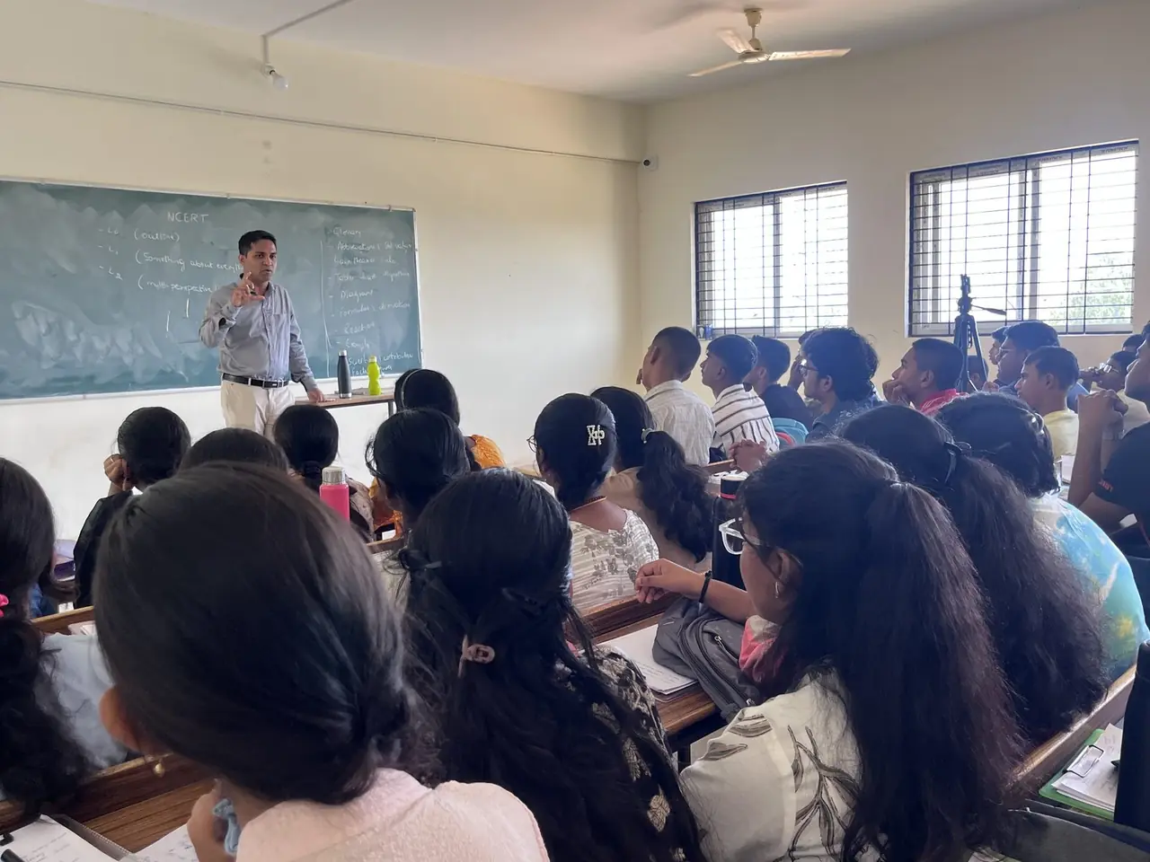 Teacher explaining a concept at the green board to a full NEET class